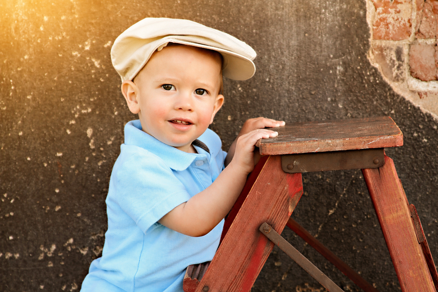A young boy wearing a light blue polo shirt and a beige cap, standing next to a wooden ladder, with a textured wall in the background, illuminated by warm sunlight.