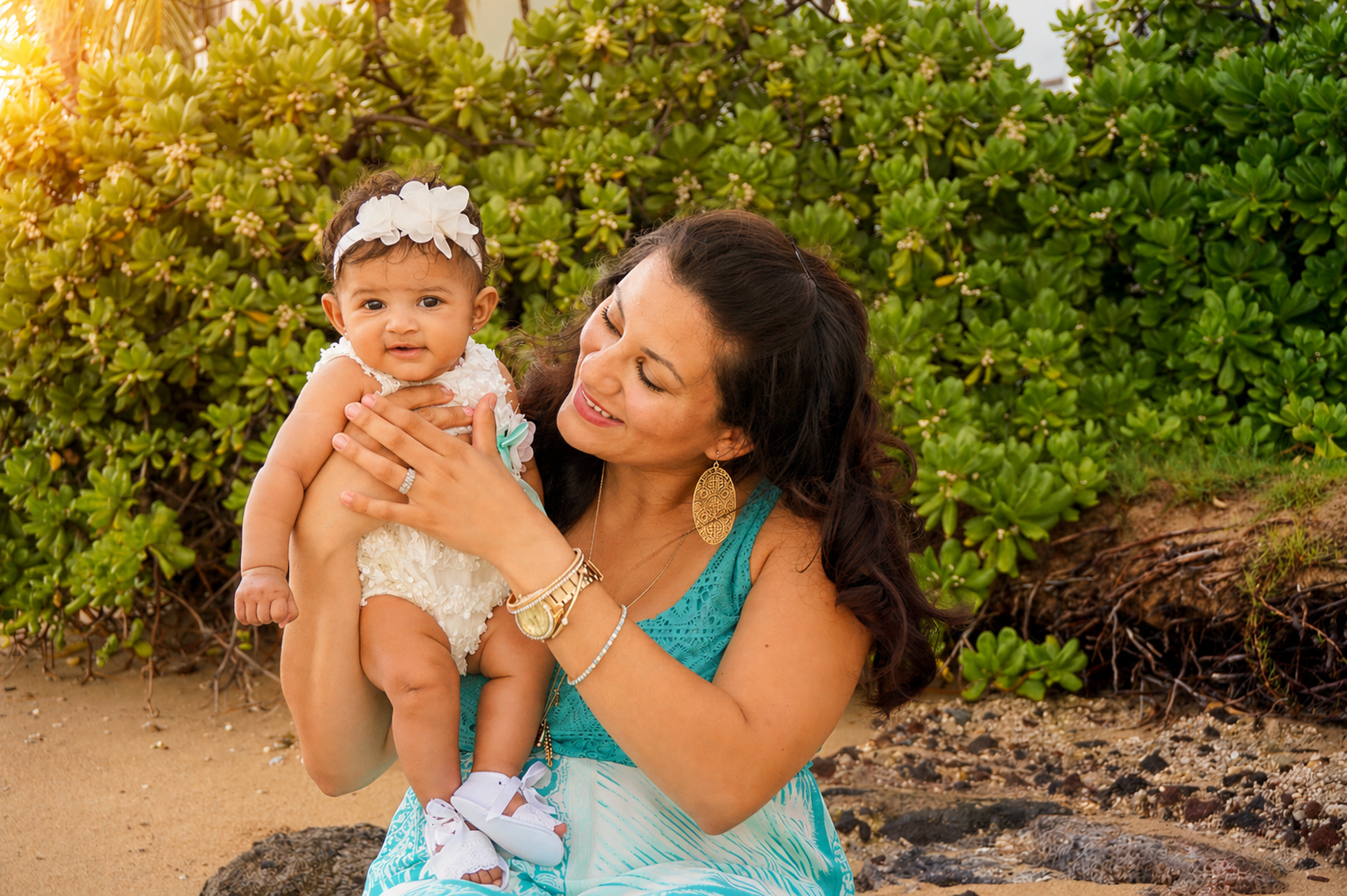 A woman holding a young girl by the beach, both smiling. The woman is wearing large earrings and a turquoise dress, while the girl is in a cream-colored dress with a floral headband. The background features green bushes and sandy ground.