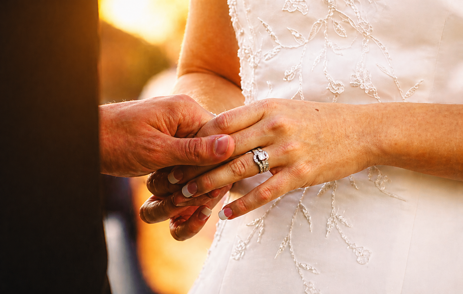 Close-up of a bride and groom holding hands during their wedding ceremony, with the bride wearing a diamond ring and an embroidered wedding dress, against a warm sunset background.