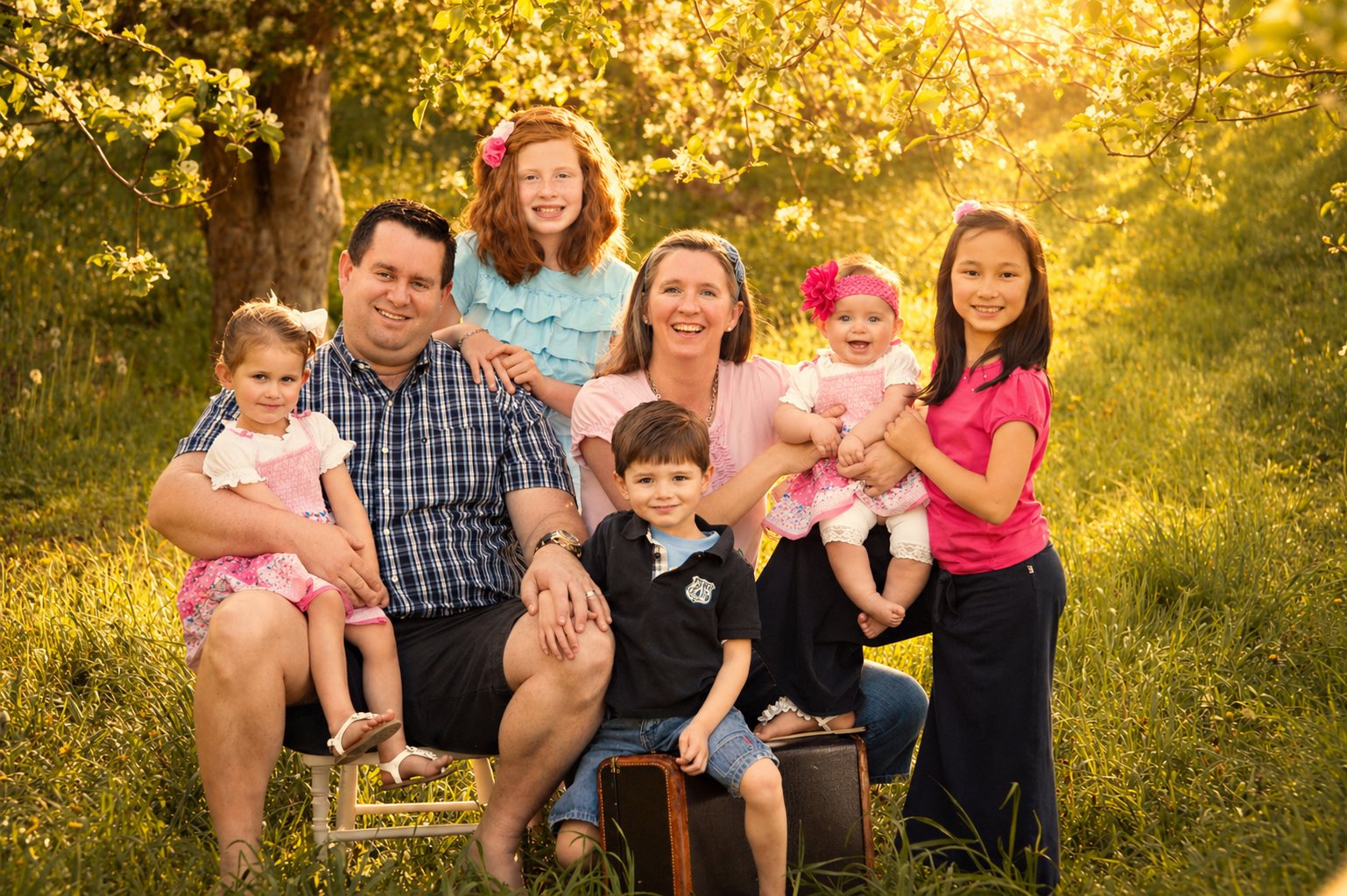 A large family of seven people, including adults and children, gathered outdoors in a grassy area with trees and sunlight filtering through the leaves, smiling at the camera.