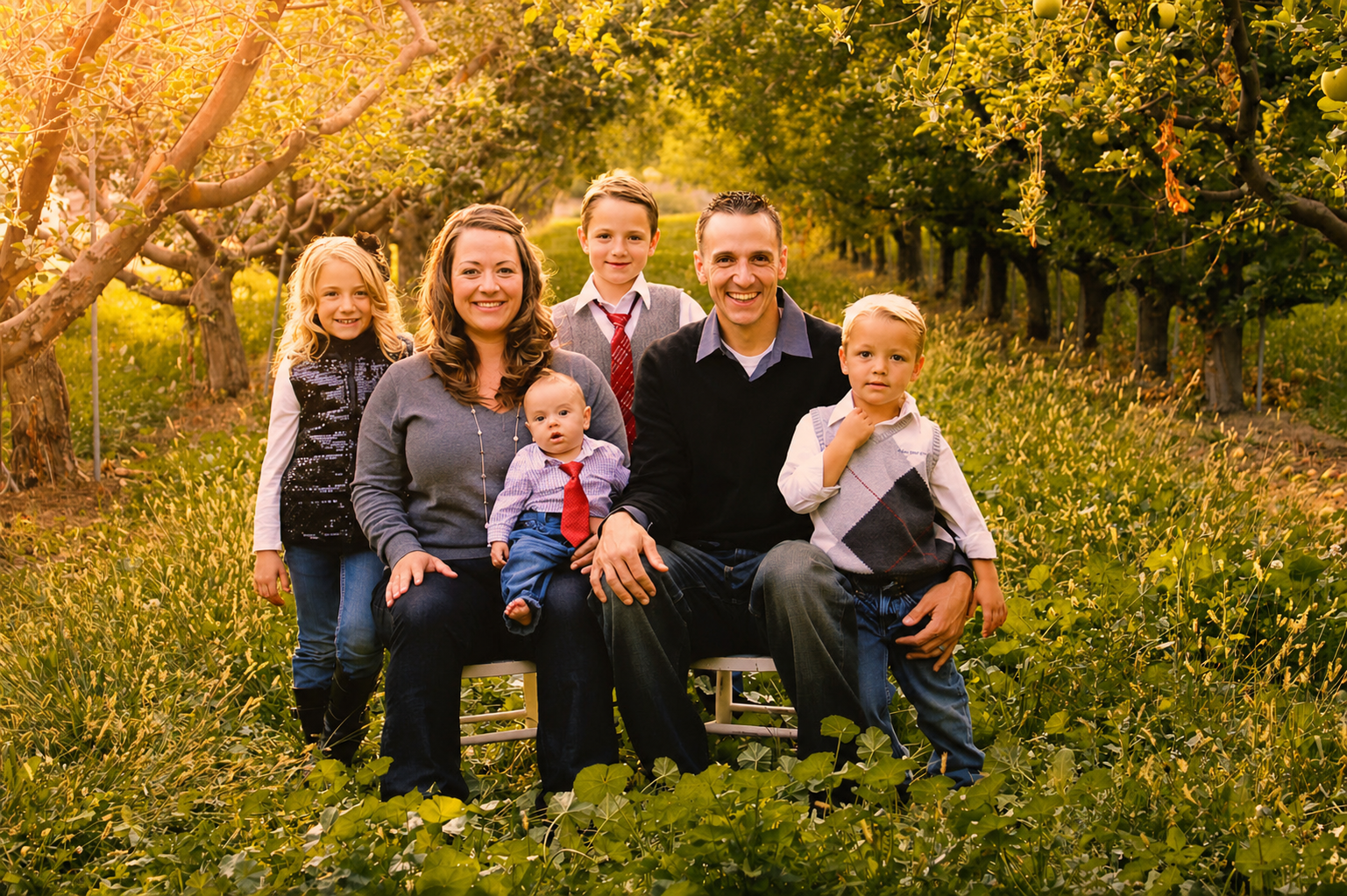 A family of seven posing in an orchard with sunlight filtering through the trees, including two adults, three children, and two babies, all dressed in casual and semi-formal clothing.