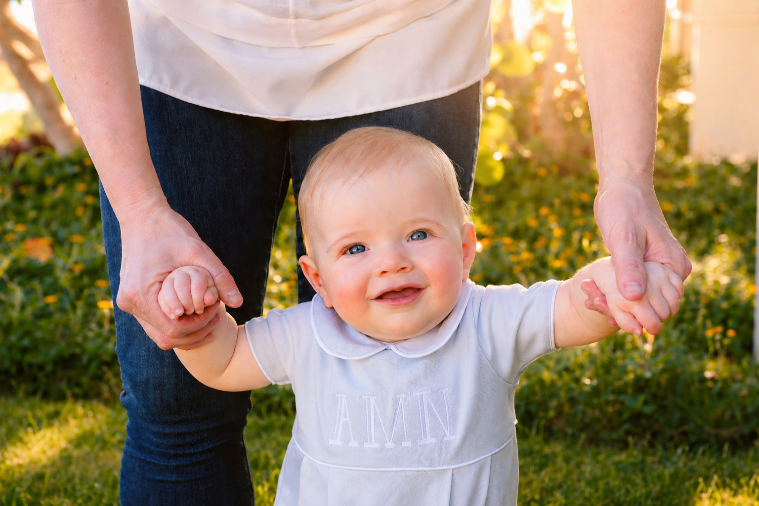 A smiling baby girl with blond hair and blue eyes, holding hands with an adult, standing outside in a garden during sunset.