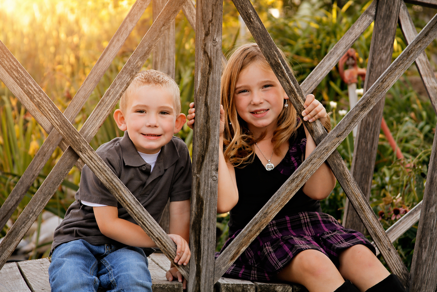 A young boy and girl sitting on a wooden bridge, smiling at the camera, surrounded by greenery and flowers during sunset.