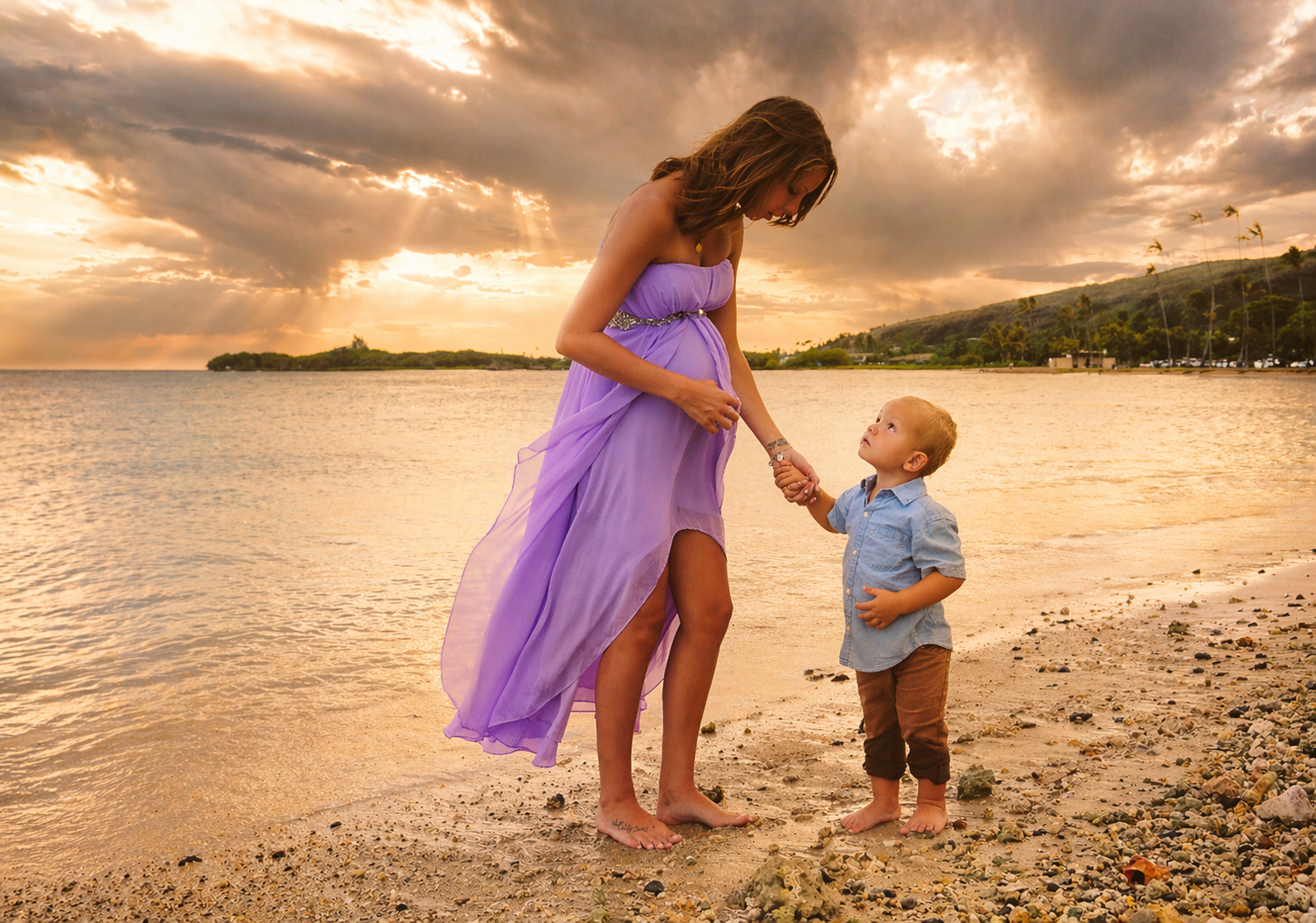 A woman wearing a flowing purple dress holding hands with a young boy in a light blue shirt and brown pants on a beach during sunset.