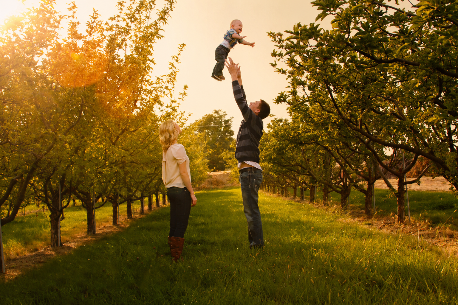 Three people in a tree-covered orchard during sunset, a man is tossing a young boy into the air while two women watch.