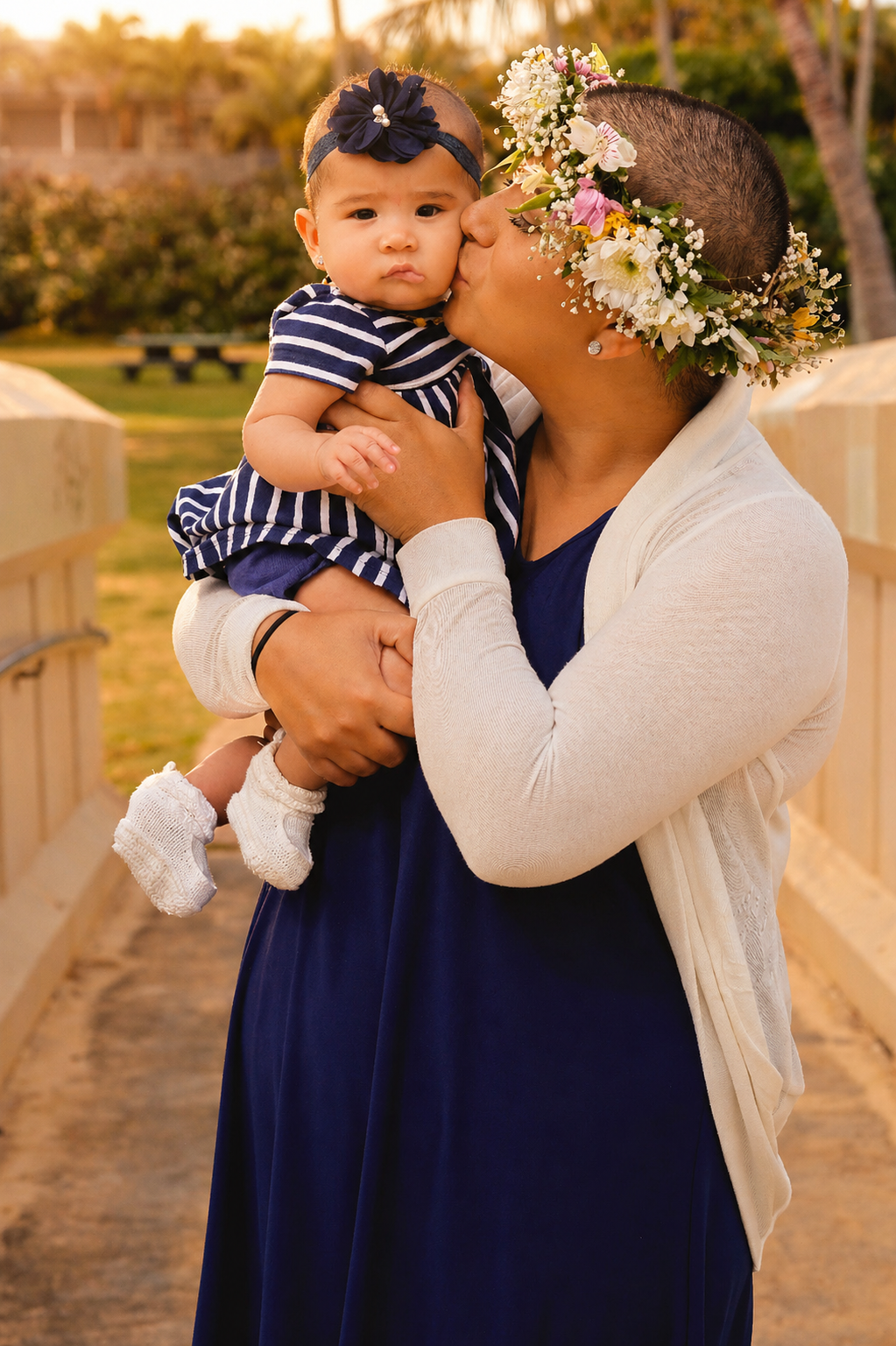 A woman with a floral crown kissing a young girl on the cheek outdoors during sunset. The girl is wearing a striped dress and white booties, and has a headband with a bow. The background shows a park with trees and benches.