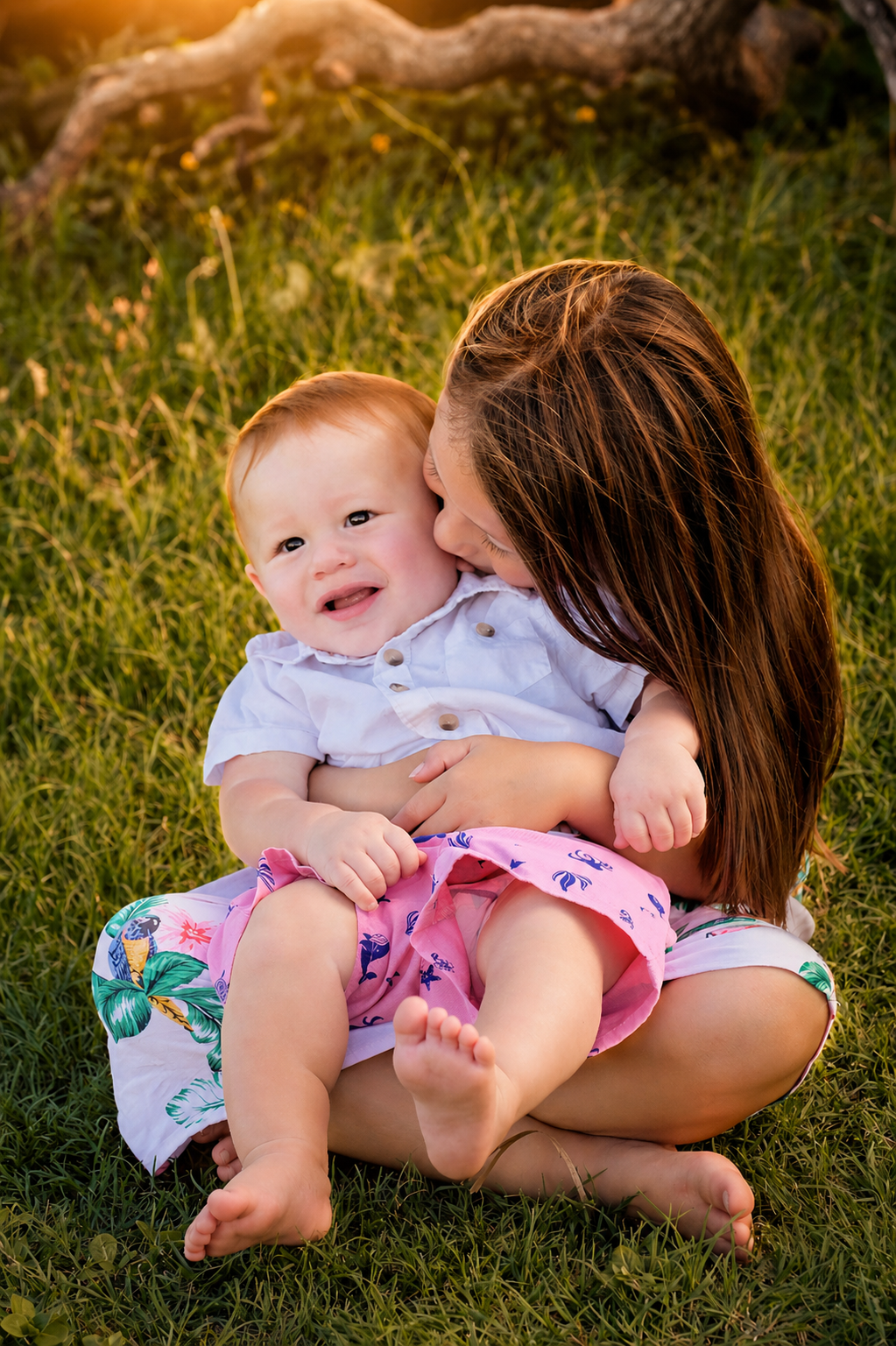A young woman with long brown hair is sitting on grass, holding and kissing a smiling toddler with red hair and light skin. The toddler is wearing a white shirt and pink shorts with a colorful pattern. They are outdoors during sunset, with a tree bra
