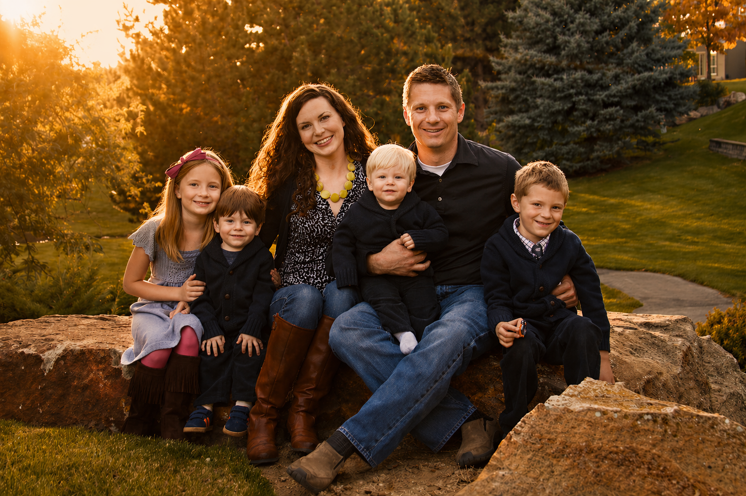 A family of seven sitting on a large rock outdoors during sunset, surrounded by trees and greenery, enjoying a warm golden hour moment.