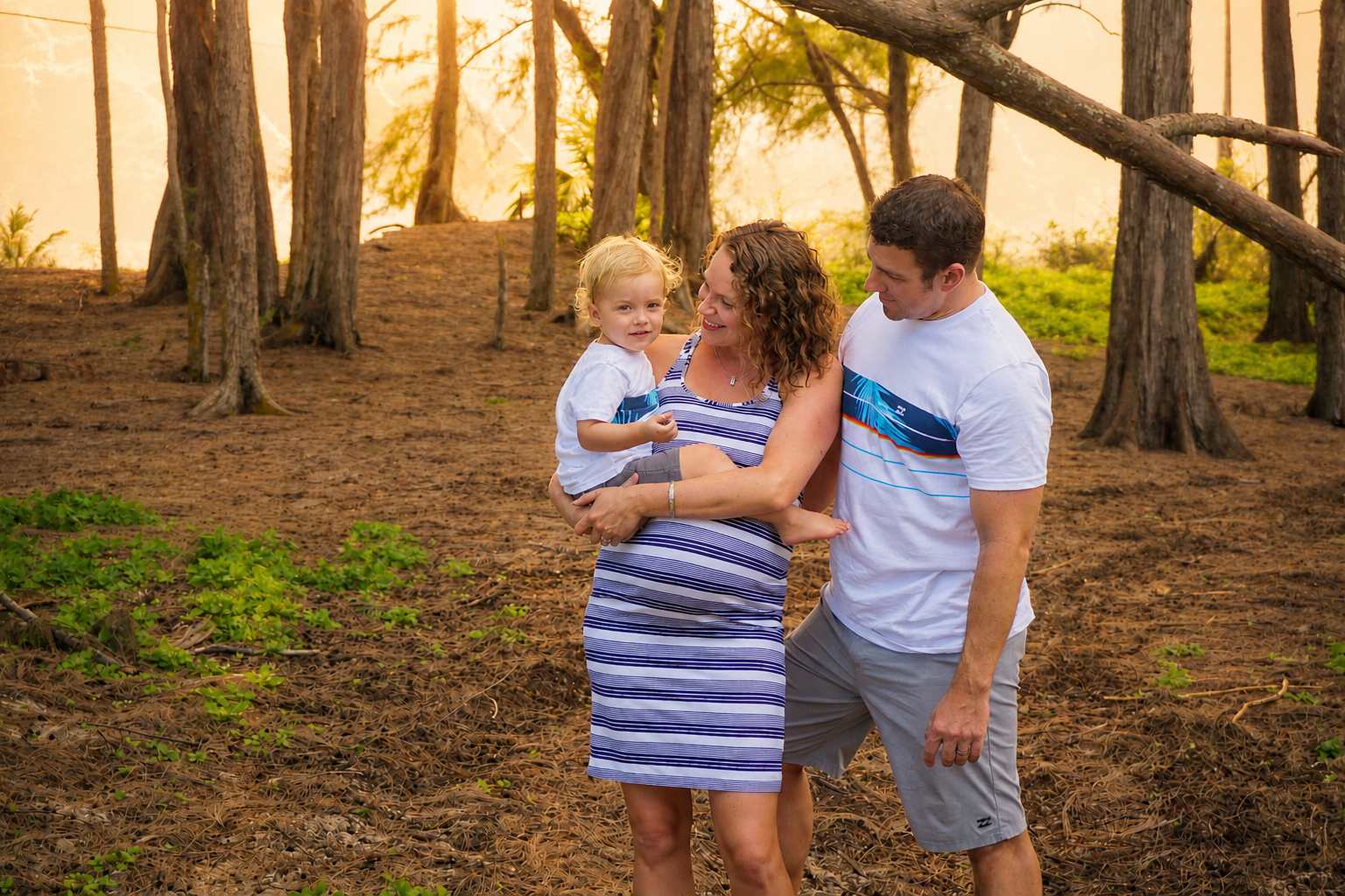 Family of three enjoying time outdoors in a forest with tall trees and green foliage, smiling and sharing a moment together.