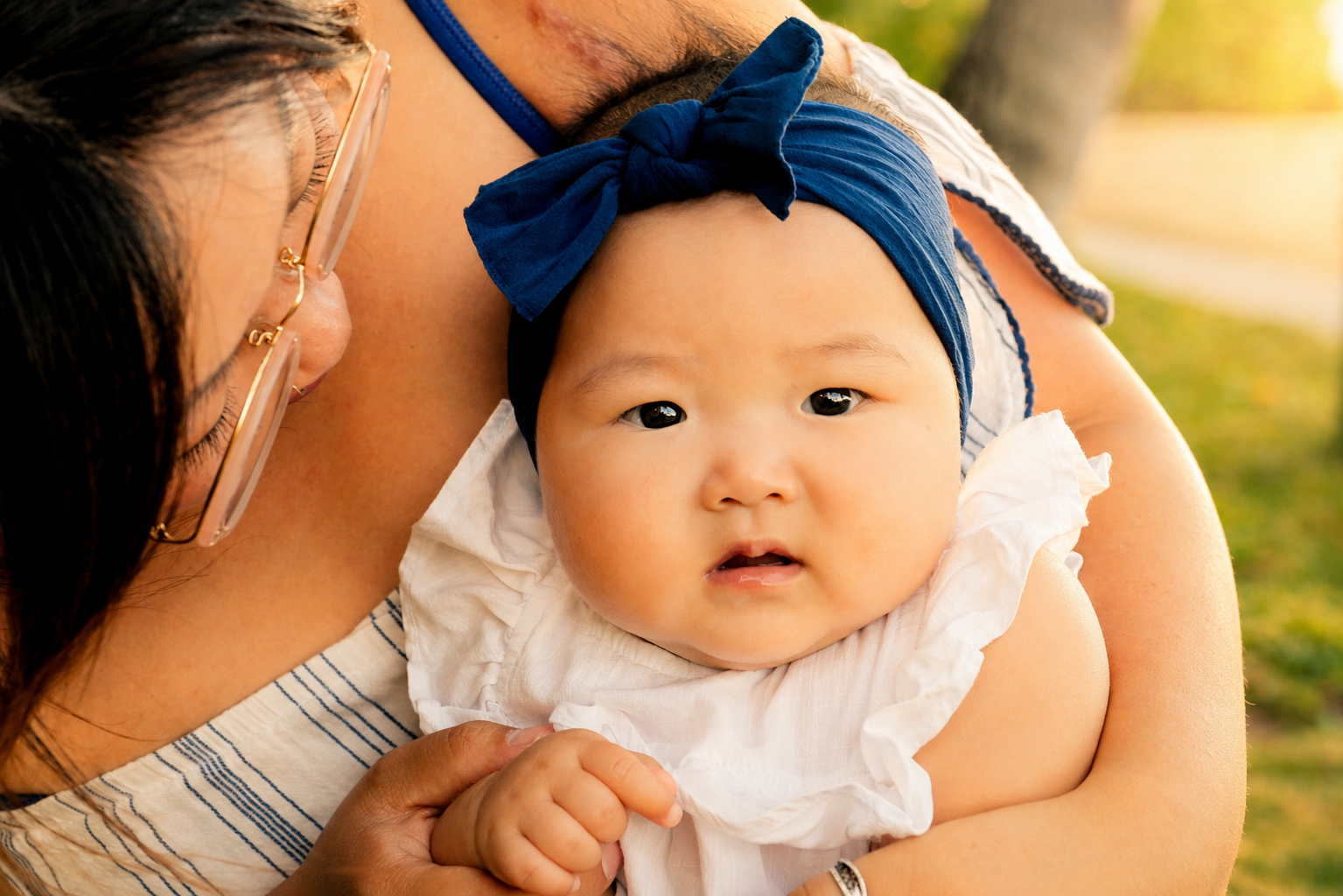 A baby with a blue headband and a white dress being held by a woman outdoors.