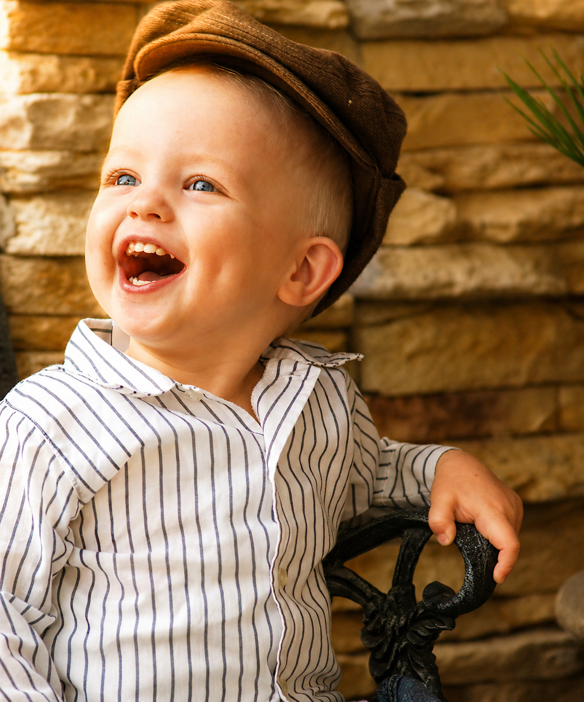 A young boy with blue eyes and blonde hair, wearing a brown cap and a striped button-up shirt, laughing happily while sitting on a black wrought iron chair in front of a wooden wall.