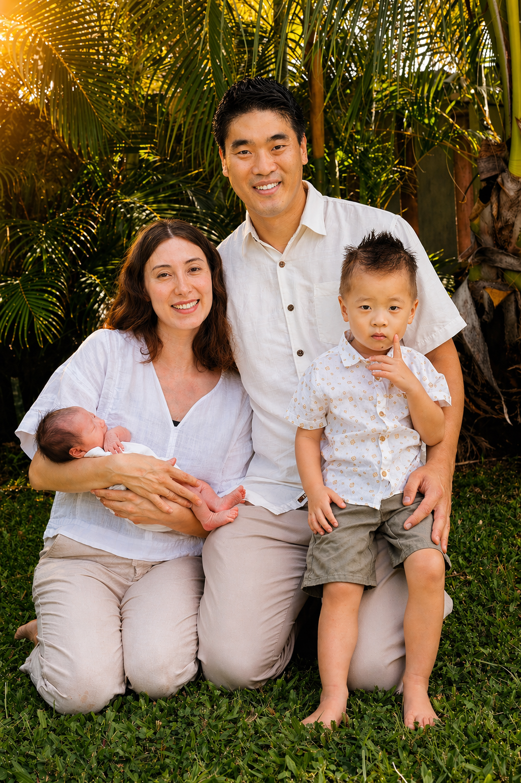 A family of four posing outdoors in a lush green tropical setting. The mother is sitting on her knees holding a newborn baby, the father is kneeling beside her with their young son standing in front of him. The family is dressed in light-colored casu