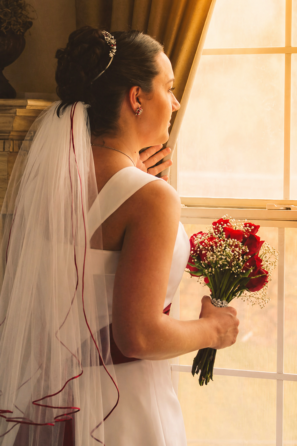 A bride in a white wedding dress holding a bouquet of red roses and white baby's breath, gazing out a window, with her hair styled up and a veil, in a warmly lit room.