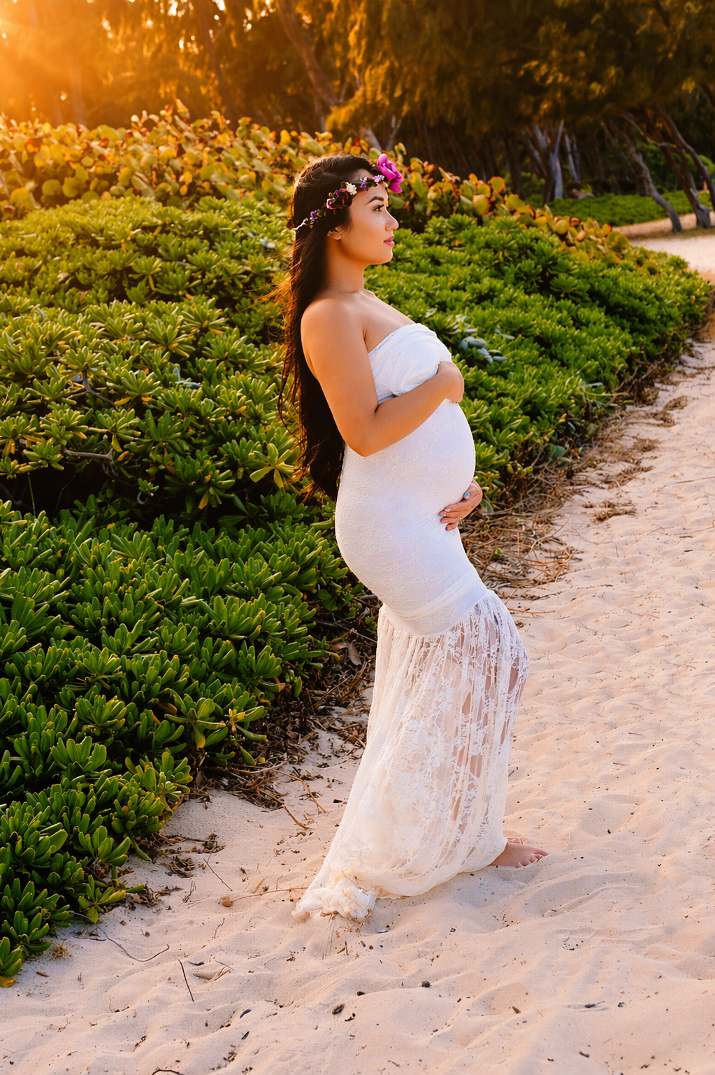 Pregnant woman in a white lace dress standing barefoot on the sandy beach near green bushes, wearing a floral headband with pink and purple flowers, during sunset.