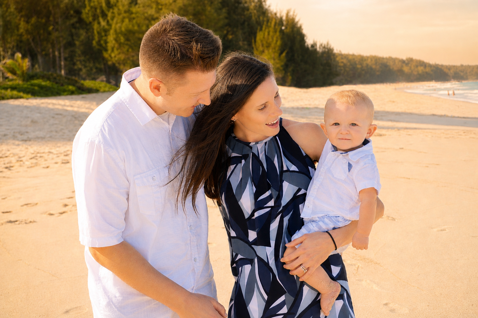A family of three, a man, woman, and a toddler boy, walking on a sandy beach at sunset with trees in the background.