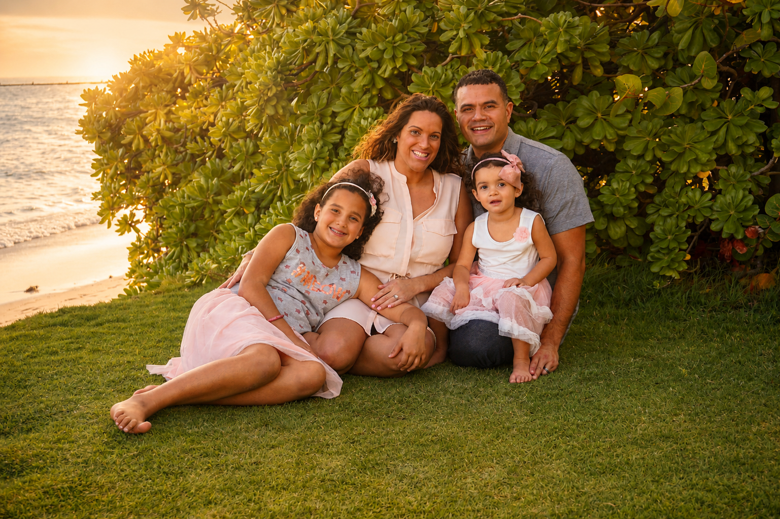 Family of four sitting on grass near the beach during sunset, smiling and enjoying the moment.