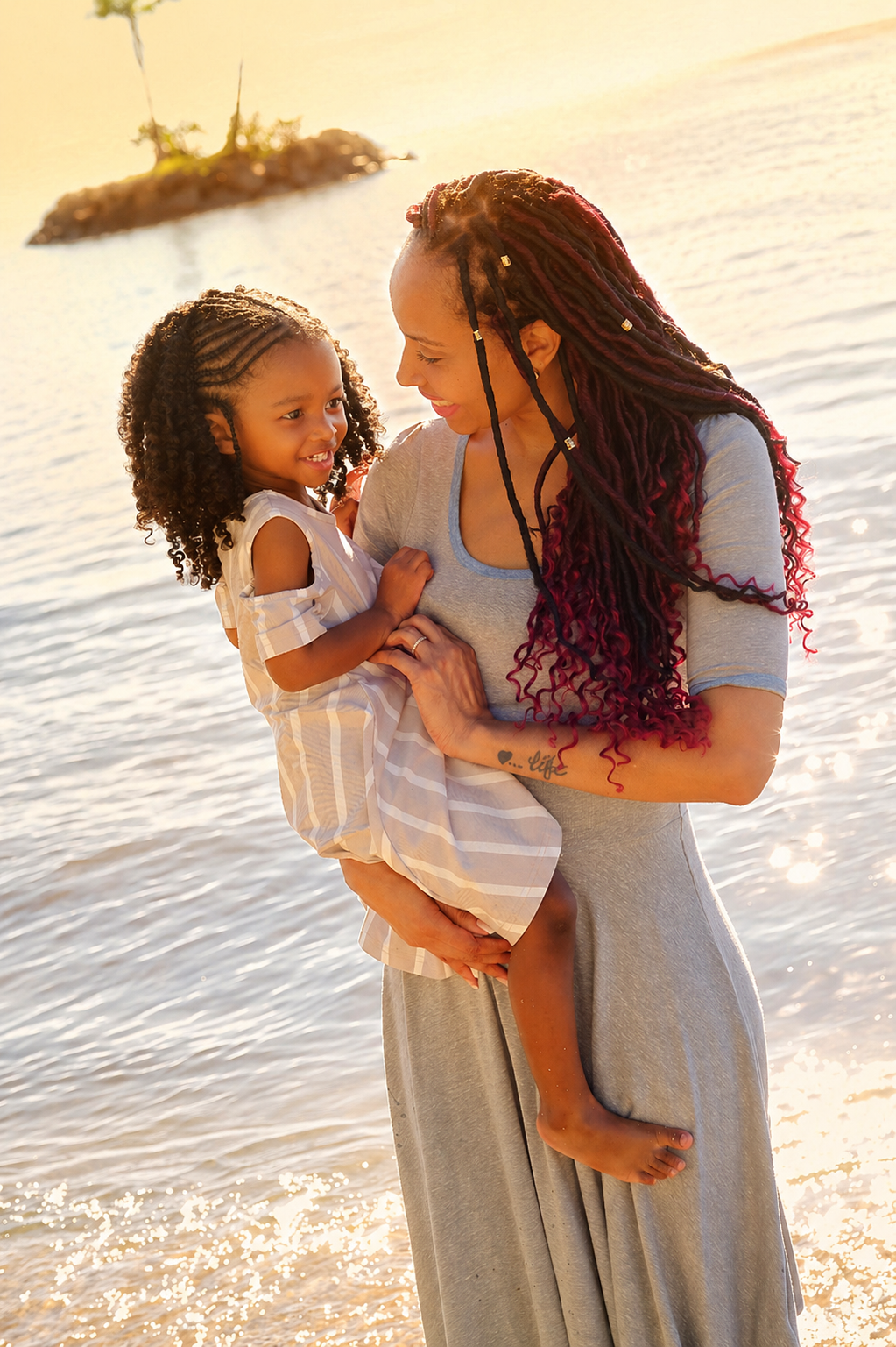 A woman holding a young girl near a body of water during sunset, both smiling and looking at each other.