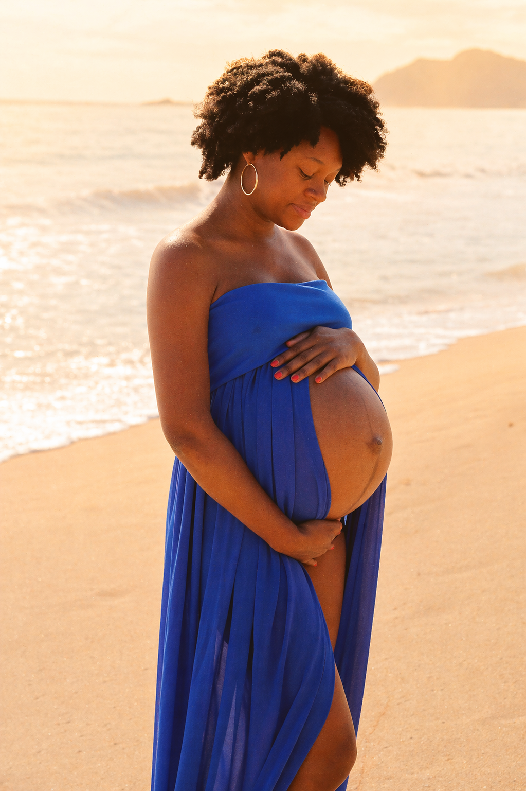 A pregnant woman in a blue dress standing on the beach during sunset, gently touching her belly and looking down.