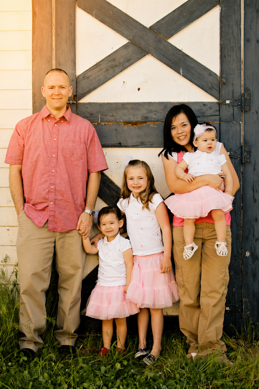 A family of six posing outdoors in front of a rustic barn door. The father on the left holds hands with a young girl, while another girl and a woman hold a toddler girl on the right. Everyone is smiling and dressed in coordinated white and pink outfi