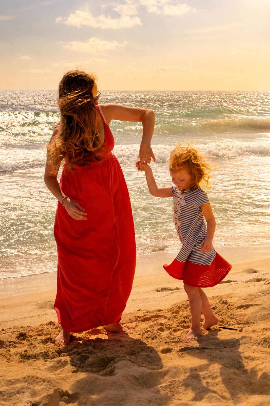 A woman and a young girl holding hands on a beach with the ocean in the background, both with curly red hair, one wearing a red dress and the other in a striped dress, during sunset.