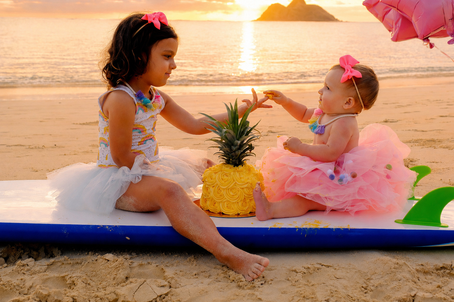 Two young girls at the beach during sunset, sitting on a surfboard with a pineapple-shaped cake between them. The older girl has dark hair and a pink bow, while the younger girl has light hair with a pink bow. The younger girl is reaching for the cak