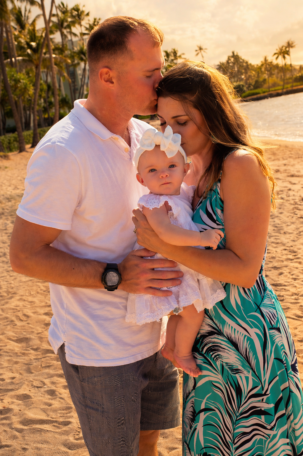 A family of three on a beach at sunset, with the father kissing the mother on the forehead, holding their baby girl who is dressed in white with a large bow in her hair.