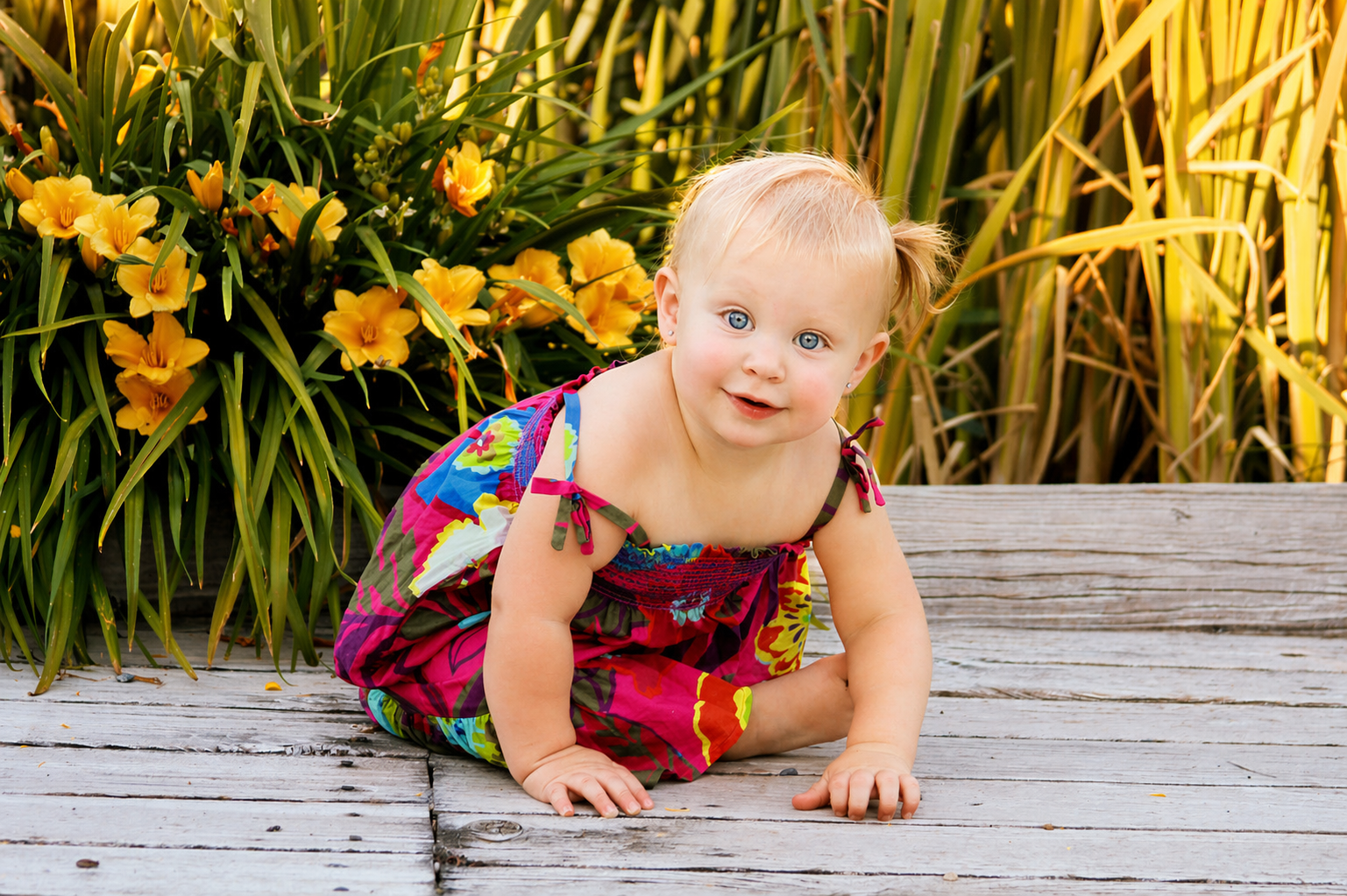 A young girl with blonde hair and blue eyes, wearing a colorful dress, crawling on a weathered wooden deck outdoors with yellow flowers and green foliage in the background.