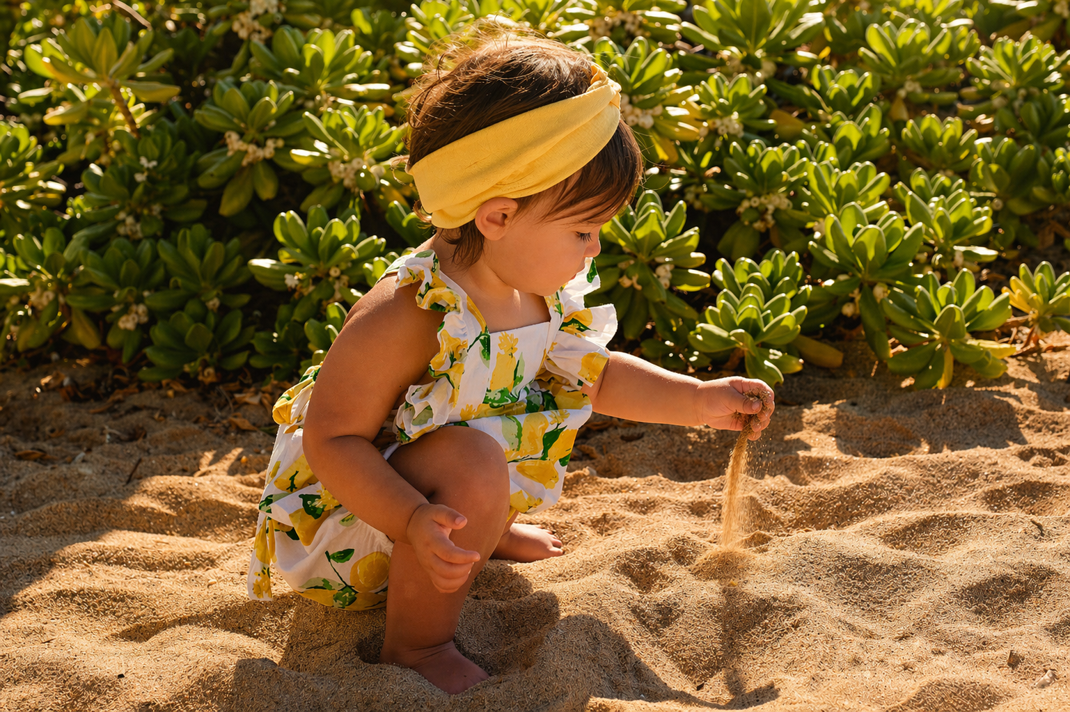 Young child squatting on sandy beach, playing with sand in front of green foliage, dressed in a floral dress and yellow headband, with sunlight illuminating scene.