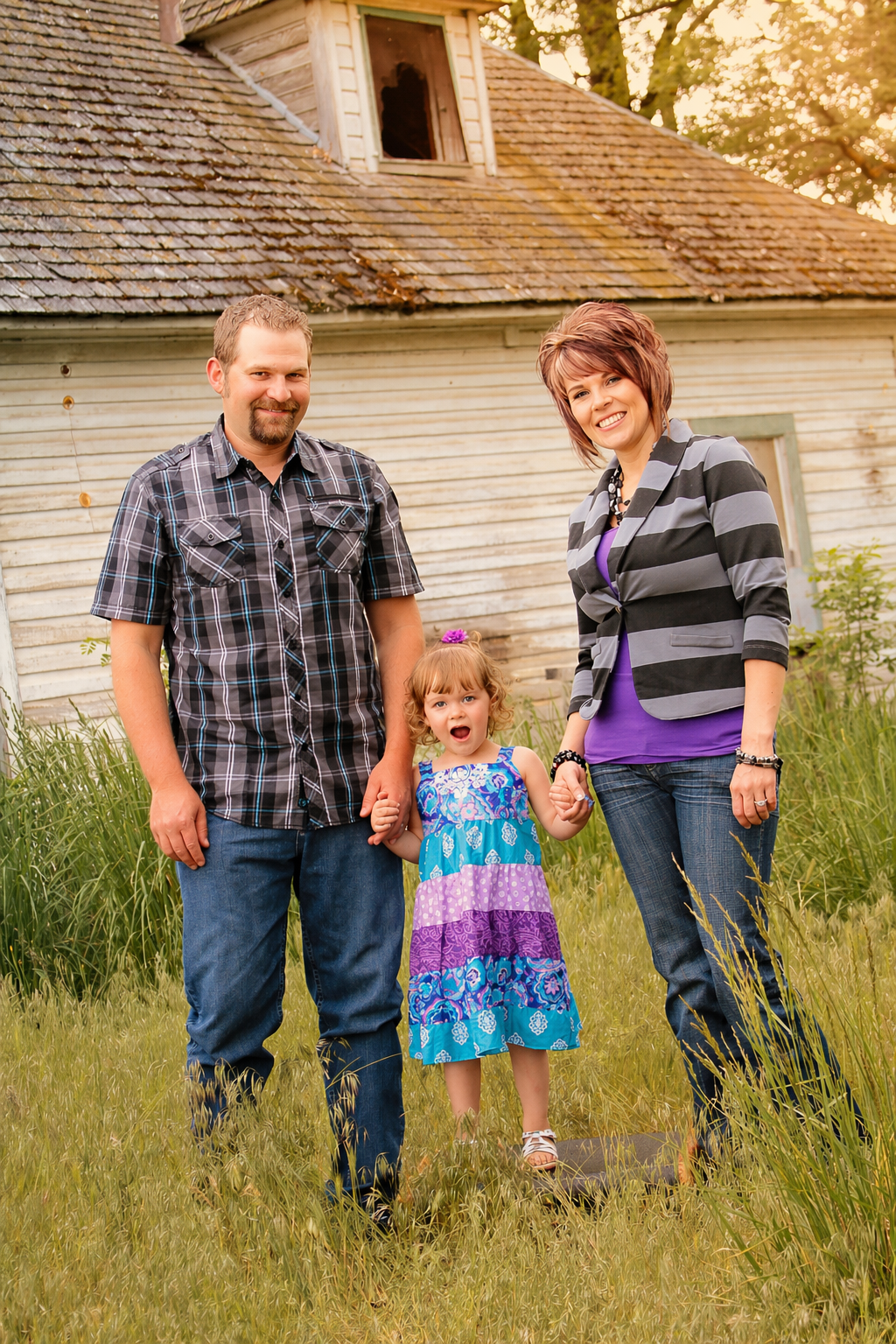 A family of three standing outdoors in front of an old wooden house, smiling and holding hands. The father has short hair and a goatee, wearing a black and gray plaid shirt. The mother has short, reddish-brown hair, wearing a striped jacket over a pu