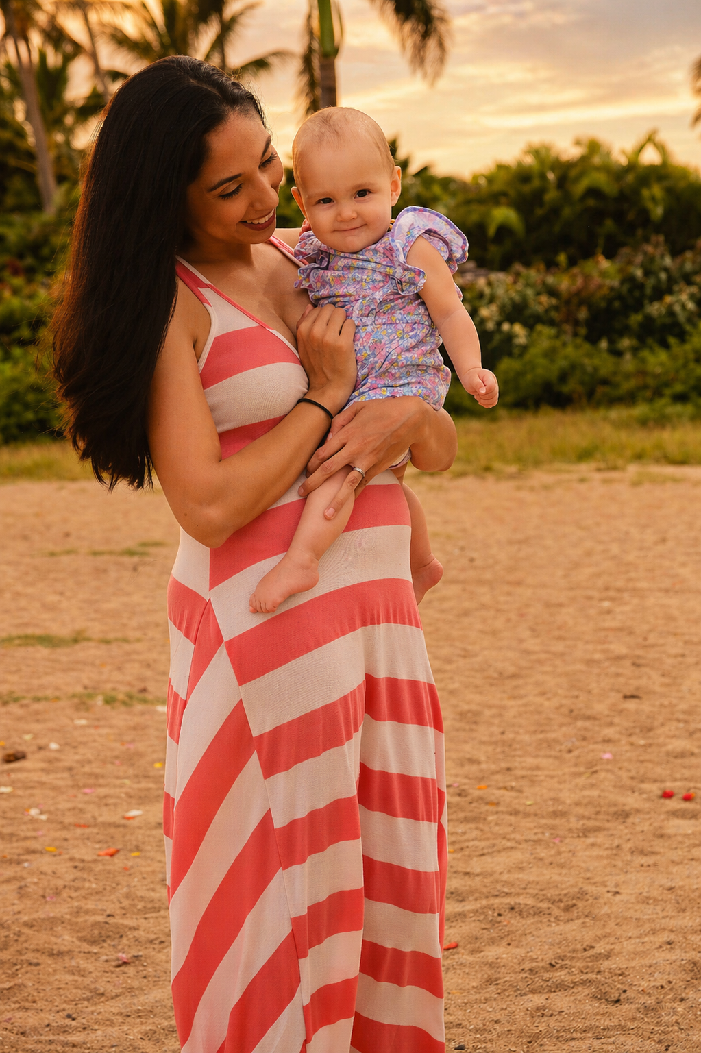 A woman with long dark hair wearing a pink and white striped maxi dress is holding a baby girl in her arms outdoors during sunset. The baby has light hair and is wearing a purple floral dress. The background features trees and a sandy area.