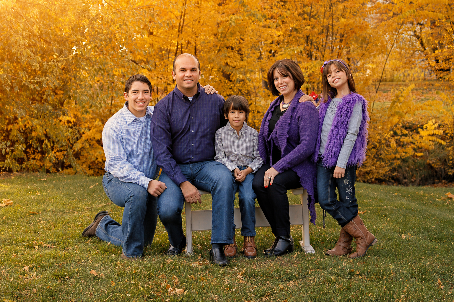 Family of six sitting on a bench outdoors during fall, with orange and yellow trees in the background. Two adults and four children are smiling and dressed in casual fall clothing.