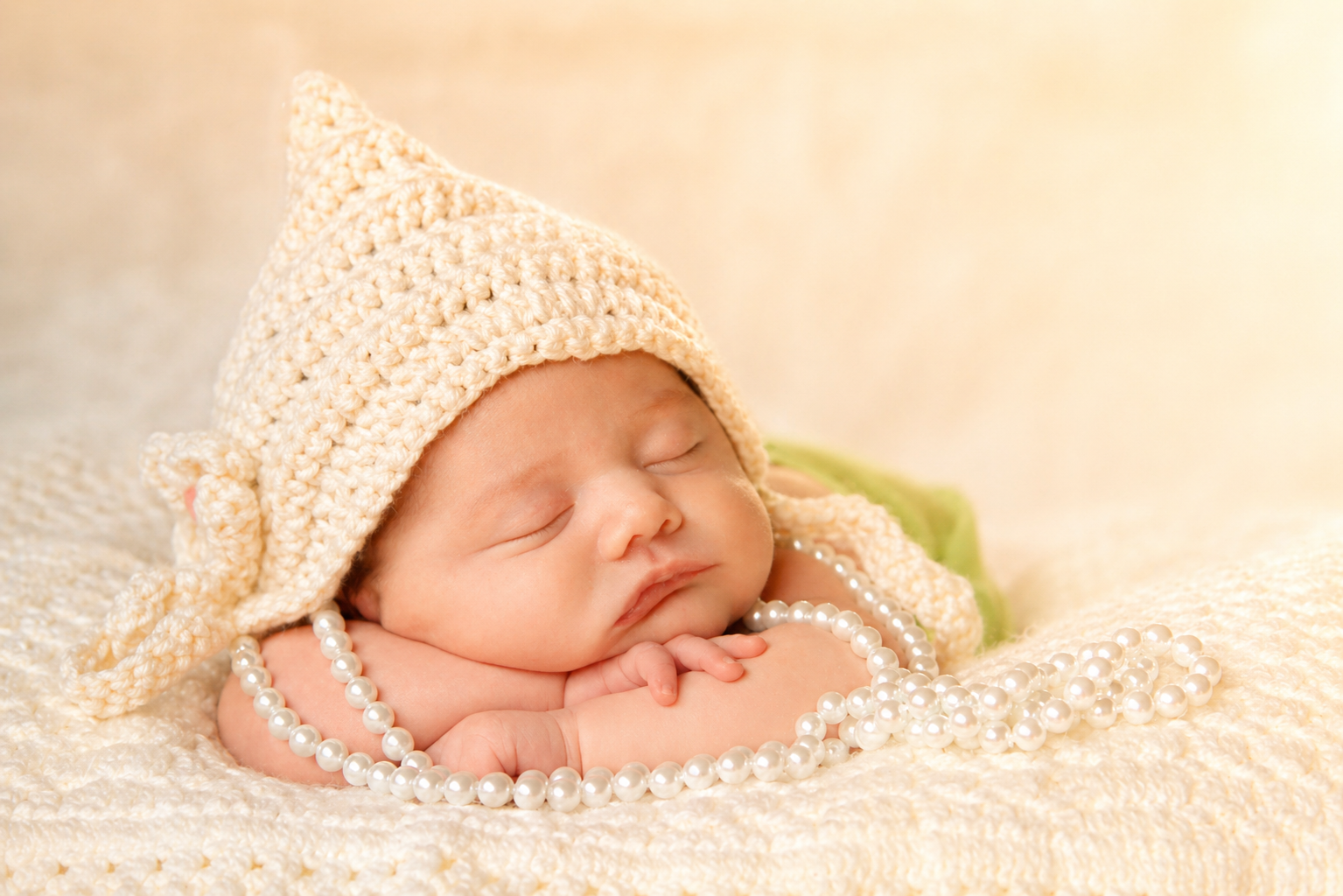 Baby sleeping on a cream blanket, wearing a cream crochet hat, with a pearl necklace, and a green blanket in the background.