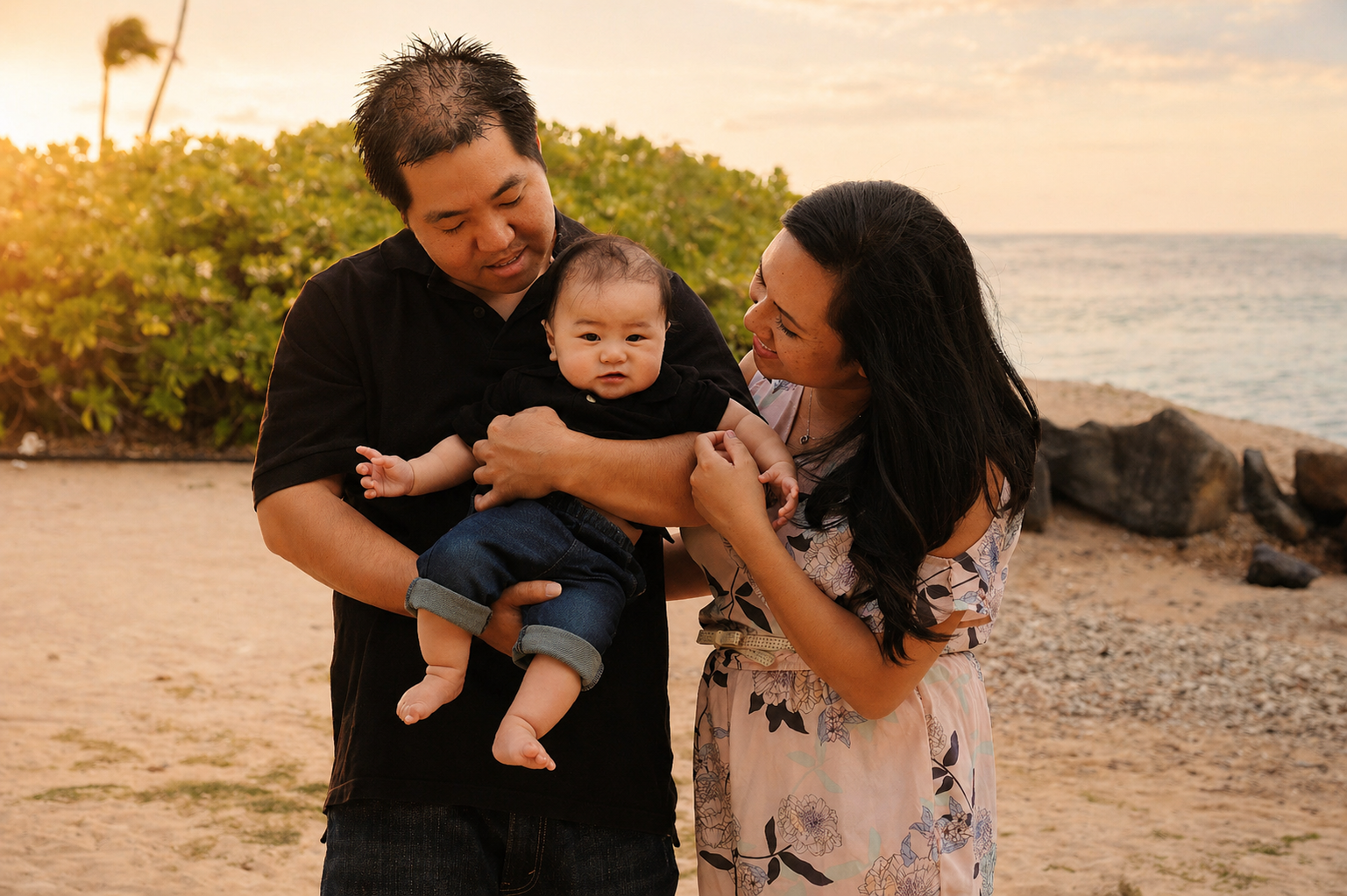 A family of three at the beach during sunset. The father is holding a baby boy, and the mother is smiling at the baby. There are rocks and bushes in the background with the ocean and sky visible.