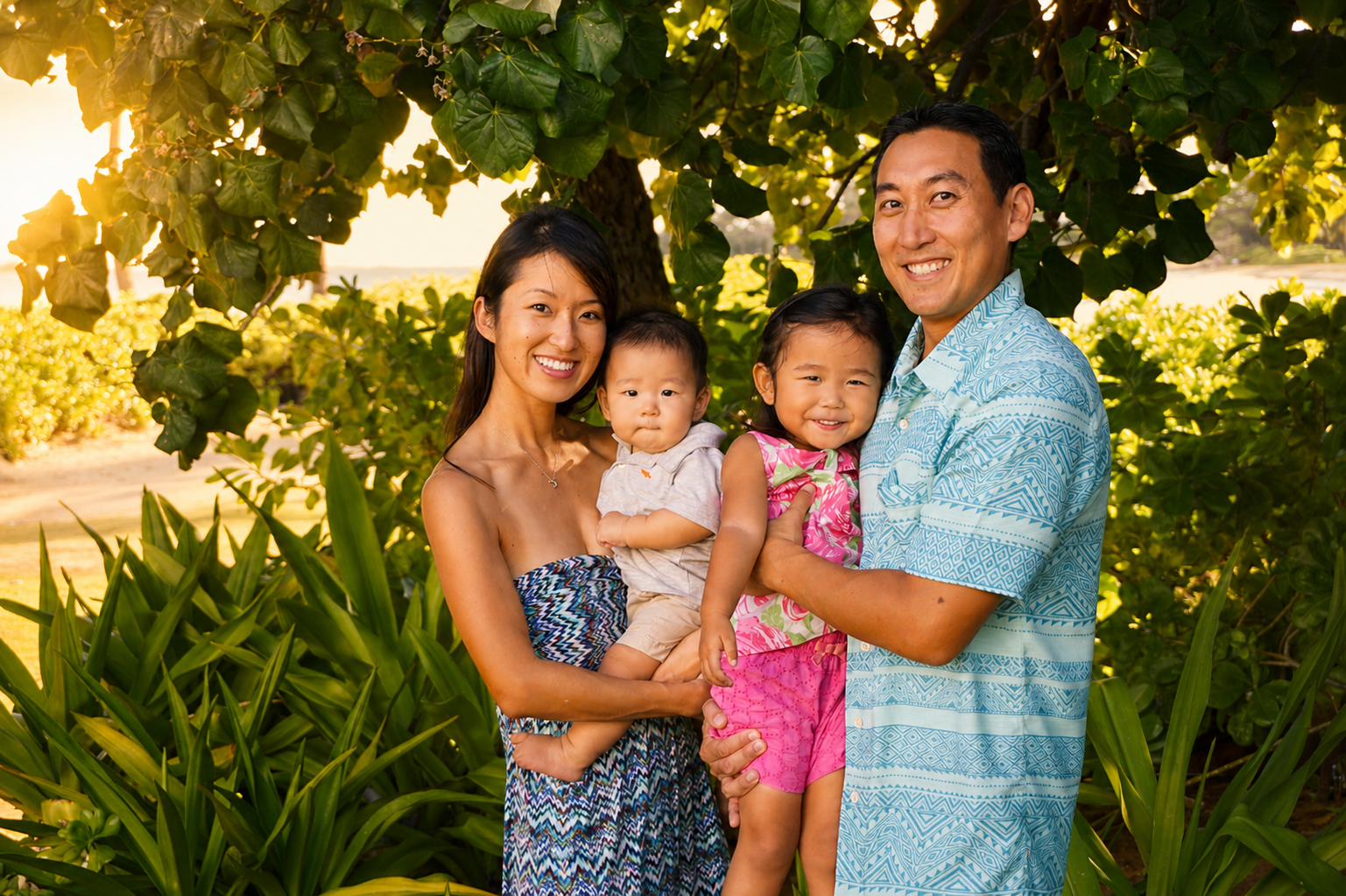 A family of four standing outdoors under a large leafy tree, smiling at the camera, with greenery and a bright sky in the background.