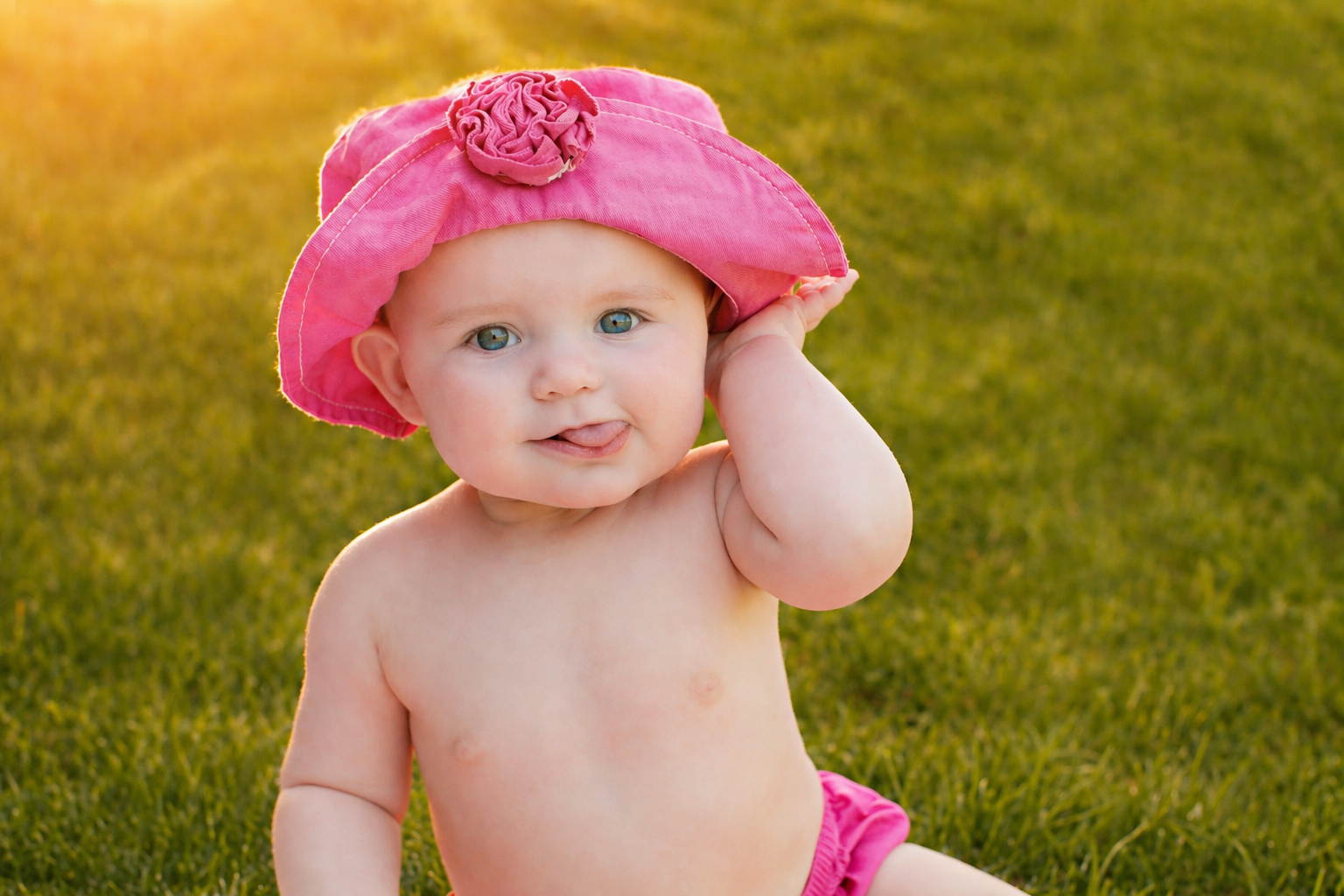 A baby girl sitting outdoors on grass, wearing a pink hat and pink bottoms, with a playful expression and sticking her tongue out.