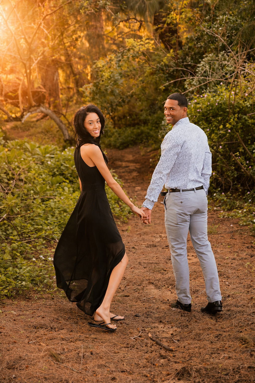 A couple holding hands and looking back, standing on a dirt path in a forest during sunset, with warm sunlight filtering through trees.
