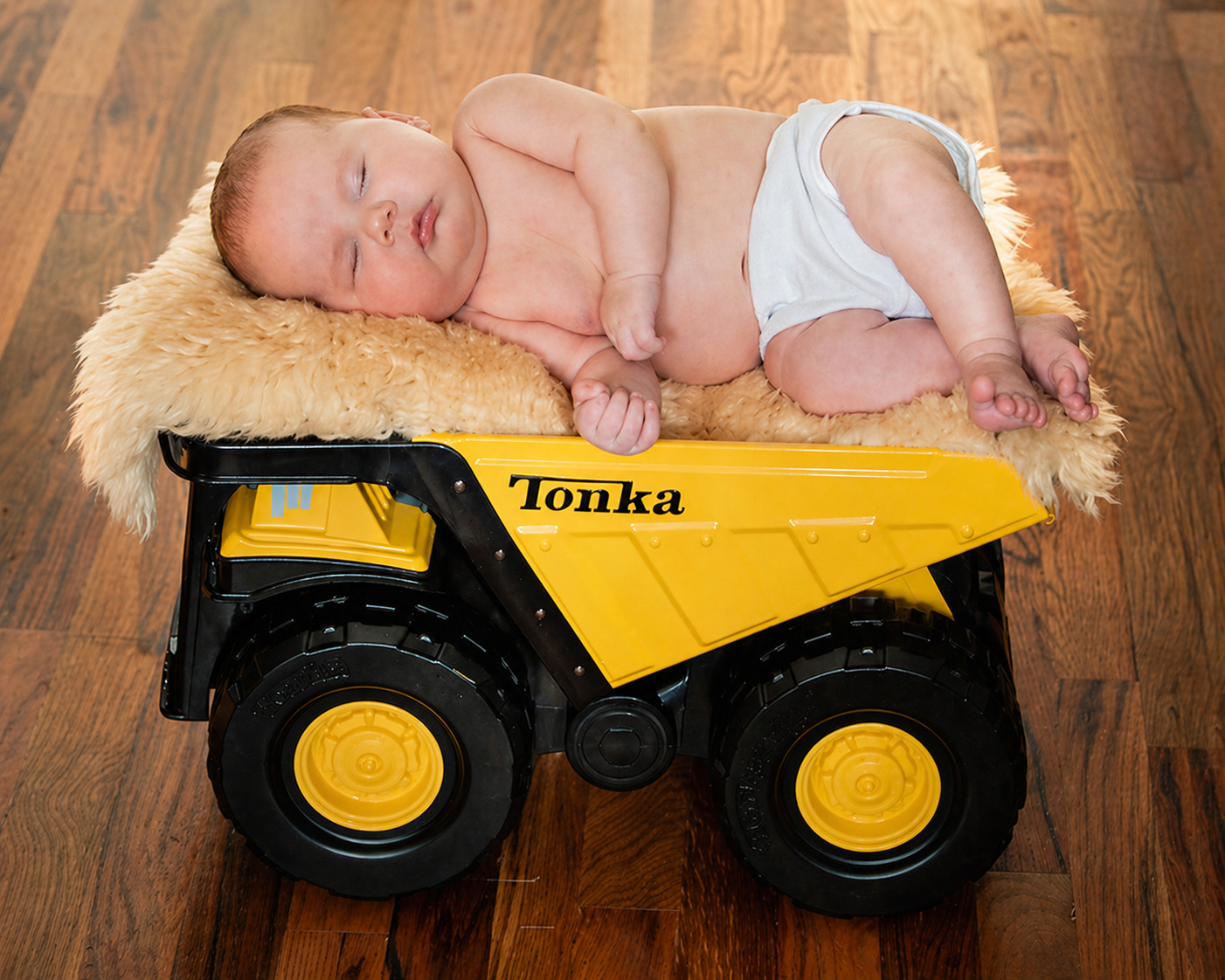 A young baby sleeping on a yellow toy dump truck with a soft fuzzy blanket on top.