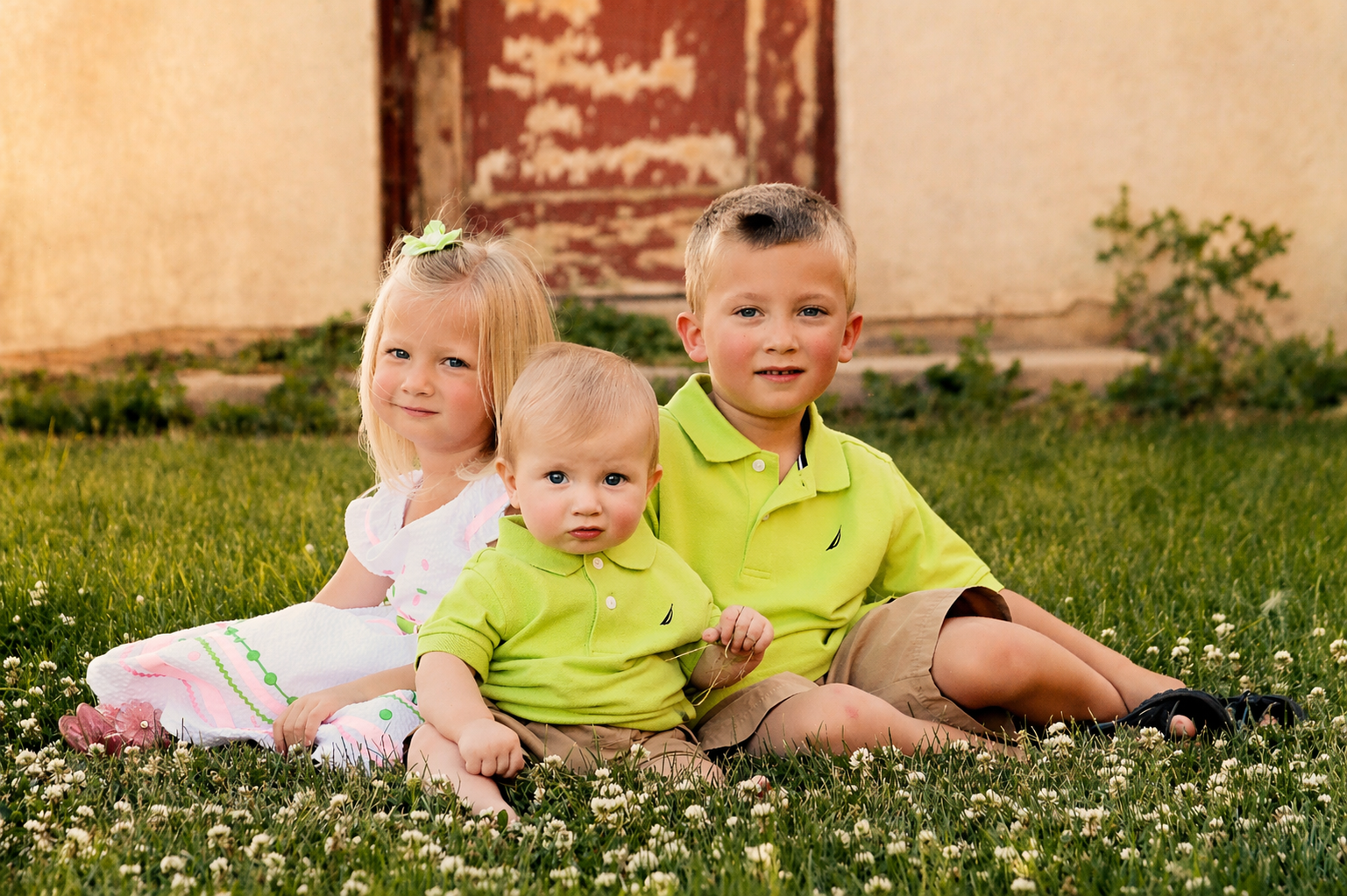 Three young children sitting on grass with white flowers, two boys and a girl, in front of a rustic building, all wearing casual summer clothes, outdoors during daytime.