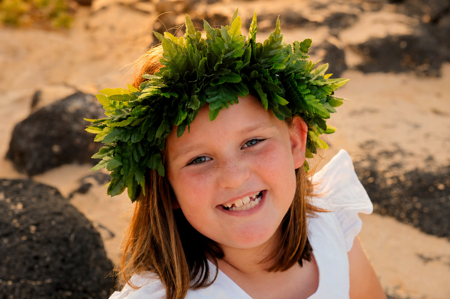 A young girl with freckles and red hair, smiling, wearing a crown made of green leaves, on a sandy beach with black volcanic rocks.