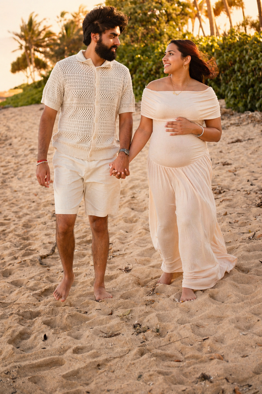 A pregnant woman and a man walking hand in hand on the beach at sunset, smiling at each other. The woman is wearing a light-colored dress and the man is wearing a light knitted shirt and shorts.