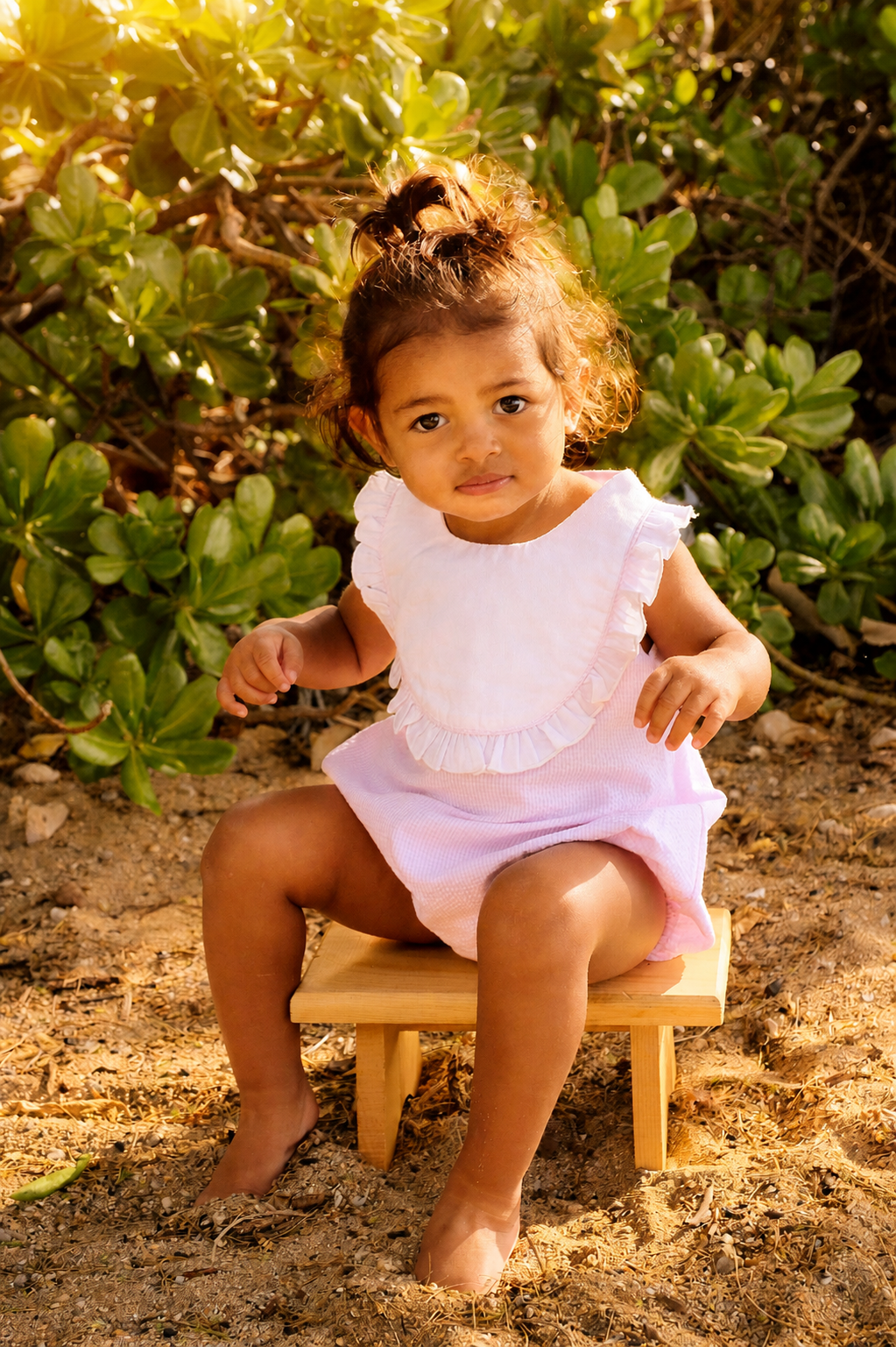 A young girl with curly hair sitting on a small wooden bench outdoors, surrounded by green bushy plants, wearing a light pink dress with ruffled collar.