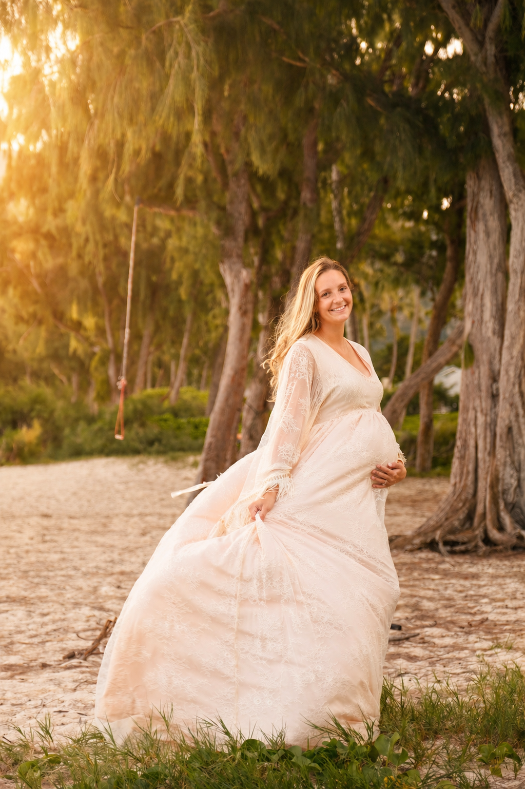 A pregnant woman in a white lace dress stands on a sandy beach, smiling and holding her belly, with trees and a swing in the background during sunset.