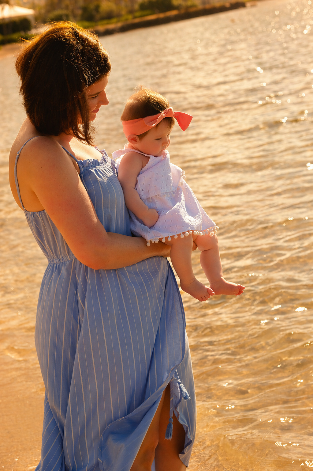 A woman in a blue striped sundress holding a young girl in a white dress with a pink headband at the beach, with water and sand visible.
