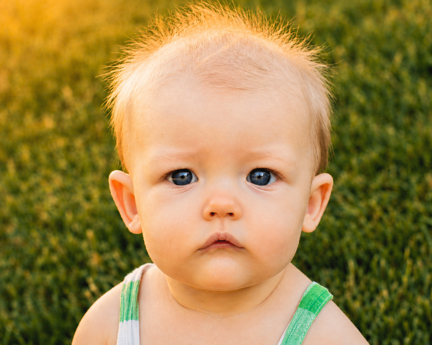 Close-up of a young child with blue eyes, light blonde hair, and wearing a green and white striped sleeveless shirt, outdoors on grass with a warm golden sunset in the background.
