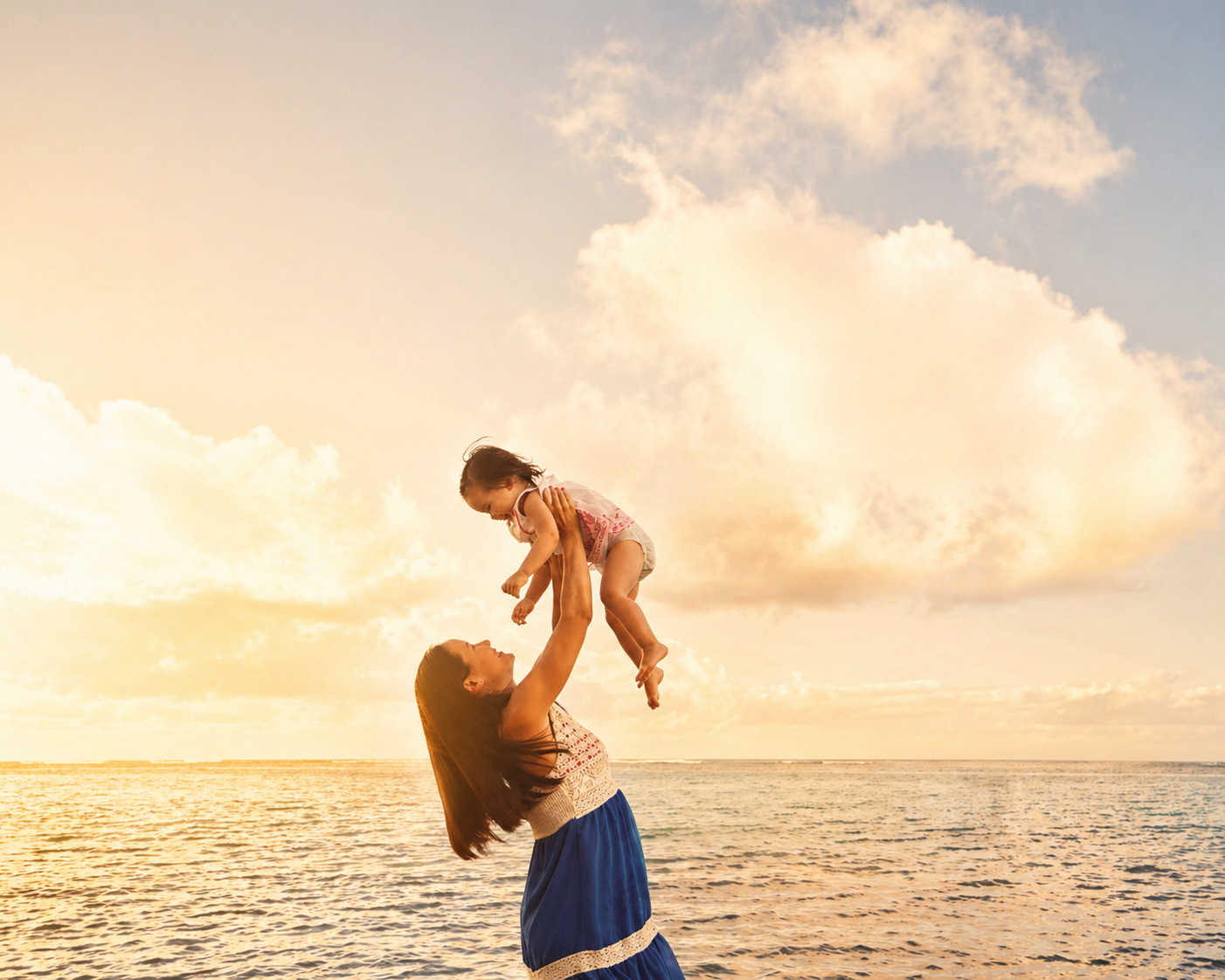 A woman playing with a young girl on the beach during sunset, with the ocean and sky with clouds in the background.