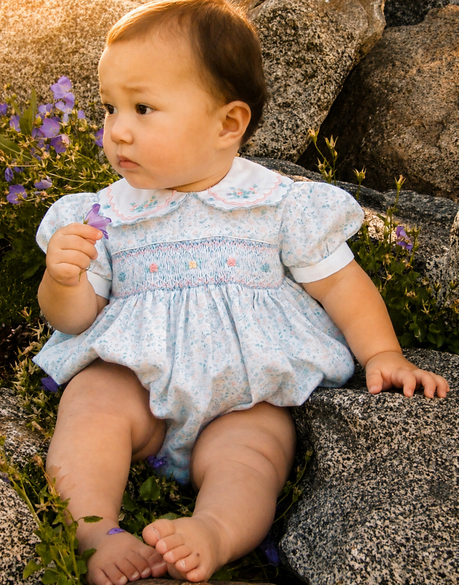 A young child, sitting outdoors on a rock, wearing a light blue dress with floral embroidery, holding a purple flower, surrounded by purple flowers and greenery, during sunset.