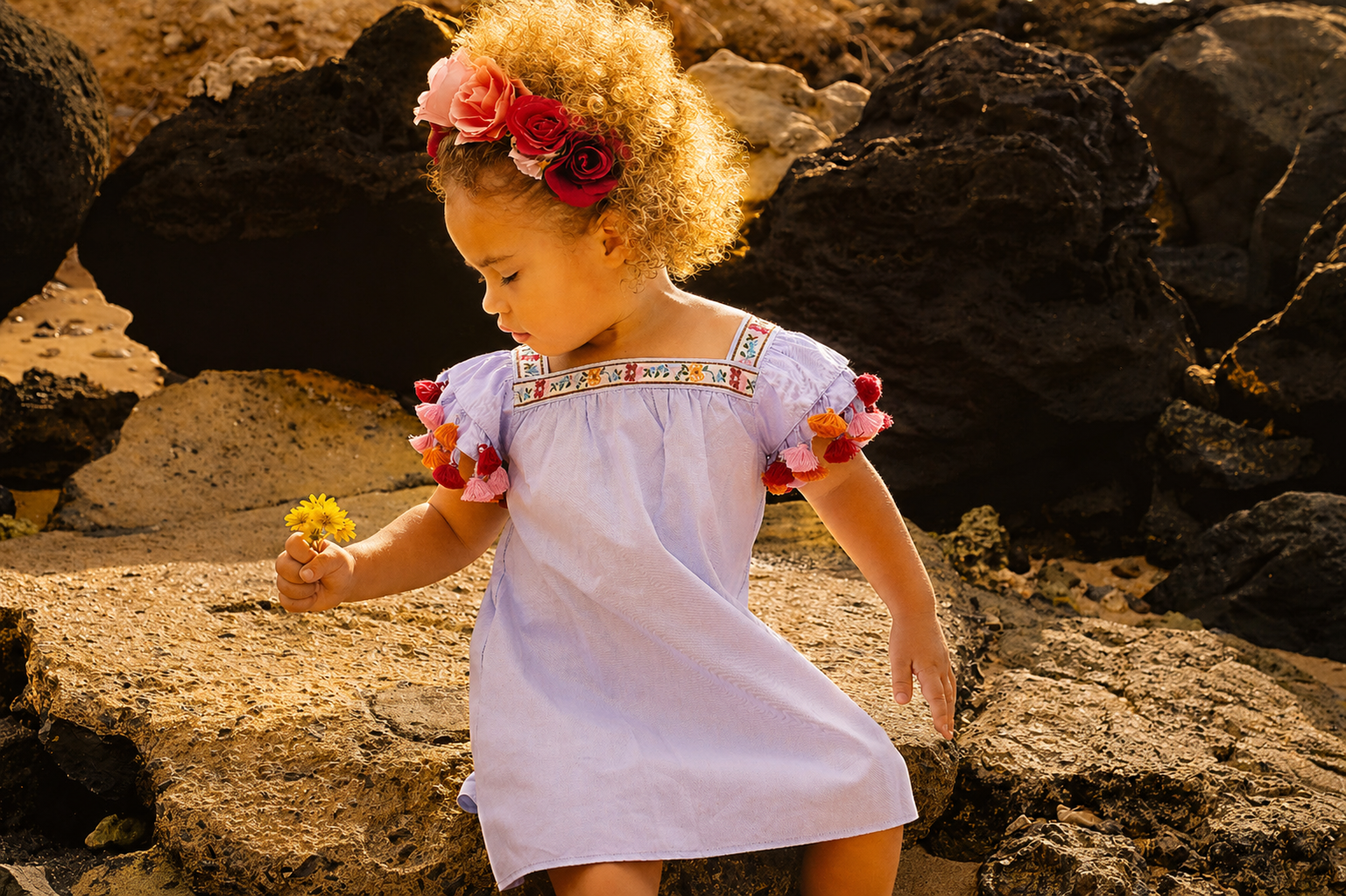 Young girl with curly blonde hair wearing a white dress with colorful embroidery and tassels, holding a yellow flower, outdoors on rocky terrain at sunset.