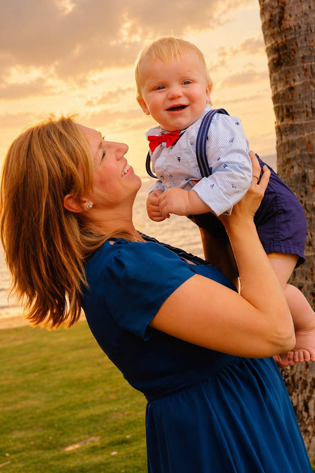 A woman with red hair smiling as she lifts a young boy with blond hair, dressed in a blue and white outfit with a red bow tie, near a palm tree at sunset.