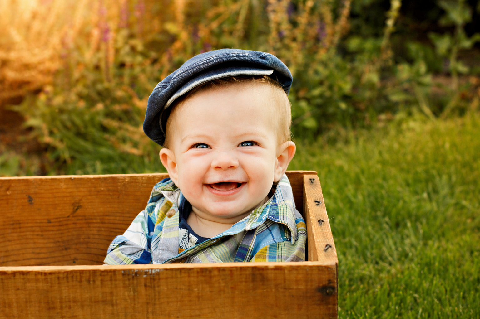 A smiling baby wearing a blue cap and a colorful striped shirt sitting in a wooden box outdoors with green grass and trees in the background.