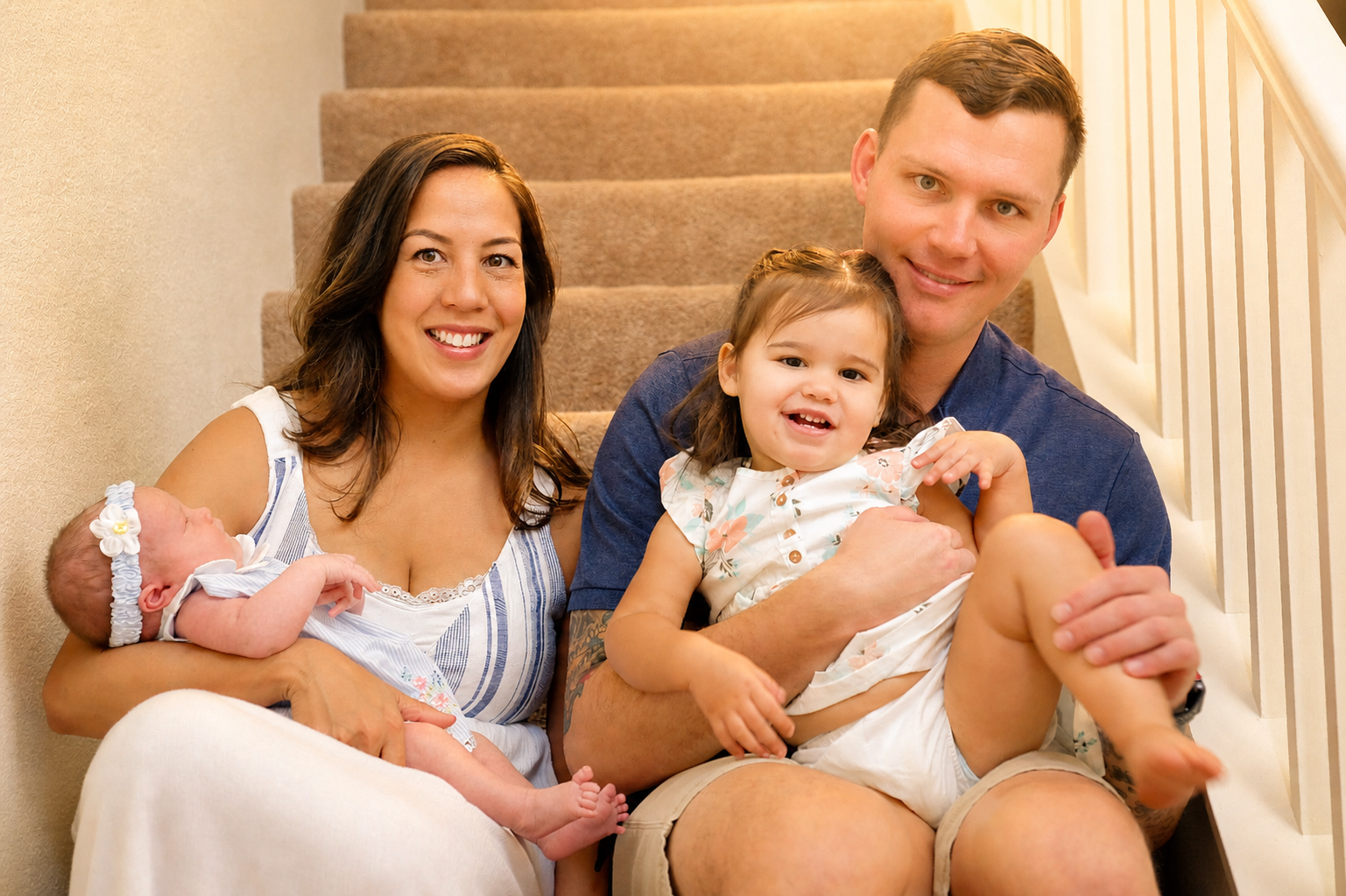 A family of four sitting on a staircase inside a home. The mother is holding a newborn baby girl, and the father is holding a toddler girl. All are smiling and looking at the camera.